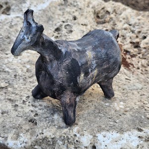 May include: A small, handcrafted sculpture of an animal, possibly a goat or sheep, with a dark, textured finish. The sculpture has a stylized form with prominent horns and is set against a natural stone background. The text "Holy Land Merchandise" is visible at the bottom.