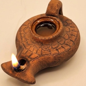 May include: A terracotta oil lamp with a lit wick, emitting a bright flame. The lamp has a circular body with a handle and a spout. The surface is textured with etched patterns. The image includes the text "Holy Land Merchandise".