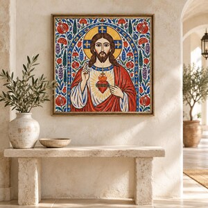 May include: A square framed wall art print depicting the Sacred Heart of Jesus. The figure is centered with a golden halo, wearing red and white robes, and holding a flaming heart. The background features blue arches, red pomegranates, and floral motifs.