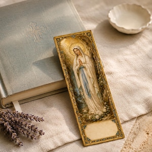 May include: A rectangular bookmark featuring a detailed illustration of the Virgin Mary in a white and blue robe with a golden halo. The bookmark has an ornate gold border and a blank rectangular space at the bottom for personalization or notes.