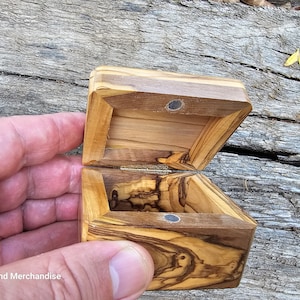 May include: A small wooden box with a hinged lid, made of olive wood with a natural wood grain pattern. The box is open and the inside is visible. The box has a magnetic closure.