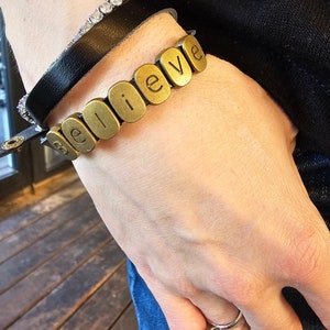 Puede incluir: Un brazalete dorado con la palabra "creer" deletreada en cuentas individuales. El brazalete se lleva en la muñeca con un brazalete de cuero negro y un brazalete de plata y cristal.