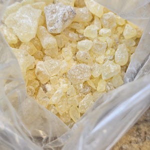 May include: A clear plastic bag filled with pale yellow, irregularly shaped resin pieces. The resin appears translucent with a slightly dusty coating. The text "Holy Land Merchandise" is visible at the bottom of the bag.