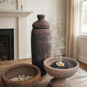 May include: A rustic wooden table displays a collection of home decor items. A tall, dark brown, lidded jar stands next to a small bowl with stones and a pedestal bowl with incense emitting smoke. The items are in a neutral-toned room.