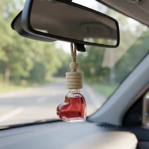 May include: A heart-shaped car air freshener filled with red liquid, hanging from a rearview mirror. The freshener has a wooden top and a beige string. The car's dashboard and a road are visible in the background.