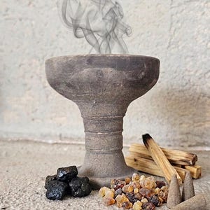 Pode incluir: Um queimador de incenso de argila cinza-escuro com uma taça larga e base de pedestal emitindo volutas de fumo. Ao lado estão pedaços de carvão preto, uma pilha de cristais de resina, vários paus de madeira clara e três cones de incenso cinzentos.