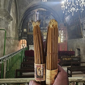 Puede incluir: Dos paquetes de velas largas y marrones, cada uno con una etiqueta, se sostienen frente a un interior de iglesia. El fondo muestra una lámpara de araña, pinturas y detalles arquitectónicos. Las etiquetas tienen texto e ilustraciones.