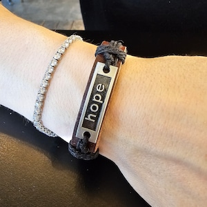 Könnte beinhalten: Ein braunes Lederarmband mit einem silbernen Anhänger, auf dem "Hoffnung" steht. Das Armband wird am Handgelenk getragen.