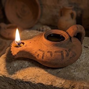 Könnte beinhalten: Eine brennende antike Öl-Lampe aus Terrakotta mit Henkel und Ausguss. Die Lampe steht auf einer Steinoberfläche, im Hintergrund ist weitere Keramik zu sehen. Die Flamme ist hell, und die Lampe hat hebräischen Text.