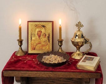 May include: A religious display featuring a gold icon of Mary and Jesus, flanked by two lit candles in brass holders. A brass censer emits smoke, and a small book rests on a red velvet cloth. A small bowl contains incense.