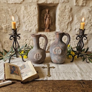 May include: A rustic display featuring two antique jugs with cross emblems, flanked by lit candles in wrought iron holders. An open book, a small cross, and a scroll are on a linen cloth, set on a wooden table against a stone wall.