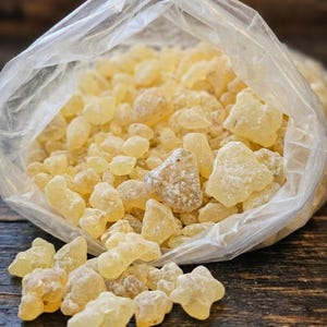May include: A clear plastic bag filled with pale yellow, irregularly shaped resin pieces. Some pieces are spilling out onto a dark wooden surface. The resin has a slightly frosted appearance, suggesting a powdery coating. The image is well-lit, highlighting the texture and color of the resin.