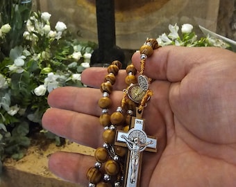 May include: A wooden rosary with a silver crucifix featuring a depiction of Jesus Christ. The crucifix has the inscription "INRI" above the figure. The rosary is held in a hand against a background of a wooden cross and flowers.