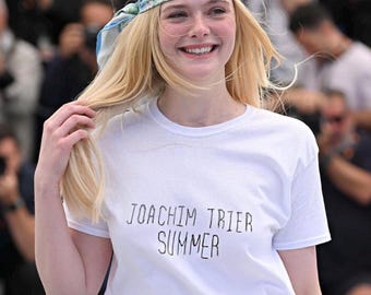 Può includere: Una maglietta bianca con la scritta "JOACHIM TRIER SUMMER" è indossata con jeans blu e una cintura marrone. Un foulard azzurro con motivi floreali è legato intorno alla testa. La cintura ha una fibbia dorata.