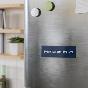May include: A navy blue rectangular magnet with the text "EVERY SECOND COUNTS" in white lettering. The magnet is attached to a stainless steel fridge door. Two round magnets, one white and one green, are also attached.