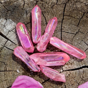 Könnte beinhalten: Rosa Aura-Quarzkristalle mit schillernden Oberflächen liegen auf einem verwitterten Holzgrund. Die Kristalle variieren in Größe und Form. Rosa Blütenblätter sind um die Kristalle herum verstreut.