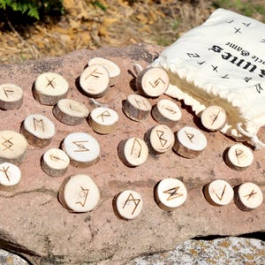 Könnte beinhalten: Ein Satz Holzrunen mit geätzten Symbolen, die auf einer Steinoberfläche angeordnet sind. Eine Leinwandtasche mit dem Text "Runes Oracle Game" ist zu sehen. Die Runen bestehen aus kleinen, runden Holzscheiben.