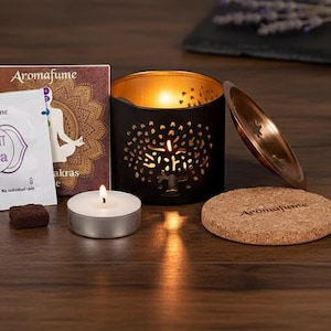 Könnte beinhalten: Aromatherapie-Set mit einem schwarzen Metall-Kerzenhalter mit Baumdesign, einem brennenden Teelicht, Räucherstäbchen-Päckchen und einem Korkuntersetzer. Das Set enthält einen kupferfarbenen Deckel und eine Box mit dem Text "Aromafume".