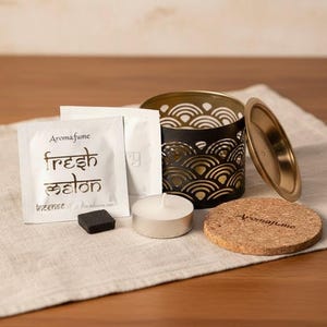 Könnte beinhalten: Aroma fume Räucherstäbchen-Set mit einem schwarzen und goldenen Metallkerzenhalter, einem Korkuntersetzer, einem Teelicht, Räucherstäbchen und Räucherstäbchen-Päckchen. Auf den Päckchen steht "fresh melon". Das Set liegt auf einem weißen Leinentuch.