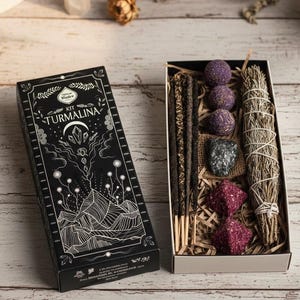 May include: A black box labeled "KIT TURMALINA" with white illustrations of mountains and crystals, next to an open box filled with incense sticks, sage, and crystals. The items are arranged on a wooden surface.
