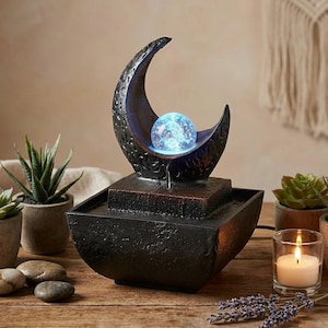 May include: A black decorative water fountain with a crescent moon design holding a glowing blue orb. The fountain sits on a square base and a bowl-shaped basin. Small potted succulents, stones, and a lit candle surround the fountain.