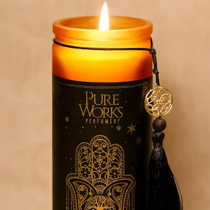 Puede incluir: Una vela negra encendida con detalles dorados y una borla negra. La vela tiene un borde dorado y presenta el texto "Pure Works Perfumery" y "Good Fortune Patchouli & Orange Manifestation Candles". Un dije dorado cuelga de la vela.
