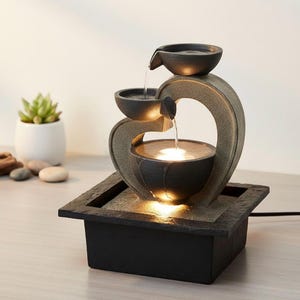 Peut inclure: Une fontaine à eau de table avec une base carrée texturée gris foncé et une structure incurvée en pierre supportant trois bols empilés. L'eau s'écoule du bol supérieur vers le bas, où le bassin est éclairé par une lumière chaude.