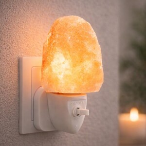 K&ouml;nnte beinhalten: Ein Himalaya-Salz-Nachtlicht, das an eine Steckdose angeschlossen ist. Die Salzlampe hat eine warme orange Farbe und strahlt ein sanftes Leuchten aus. Das Nachtlicht hat einen Ein-/Ausschalter und ist auf einem wei&szlig;en Sockel montiert.