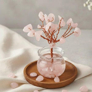 Könnte beinhalten: Dekorative Rosenquarz-Baumskulptur. Der Baum hat einen Stamm und Äste aus Kupferdraht, besetzt mit rosa Rosenquarzsteinen. Er steht in einer kleinen Glasvase mit Rosenquarz auf einem Holztablett. Mehrere lose Steine liegen verstreut.
