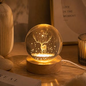 Könnte beinhalten: Eine klare Glaskugellampe mit einer lasergravierten Darstellung eines Hirsches inmitten von Sternen. Die Kugel ruht auf einem runden, hellen Holzsockel mit einem weißen Stromkabel an der Seite und strahlt ein warmes, goldenes Licht aus.