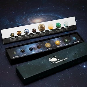 Könnte beinhalten: Ein Set aus Edelsteinplaneten in einer schwarzen Box mit dem Text "GEM PLANETS". Die Planeten sind in einer Reihe angeordnet, wobei jeder Planet eine einzigartige Farbe und Textur hat. Die Box befindet sich auf einem Hintergrund einer Galaxie.