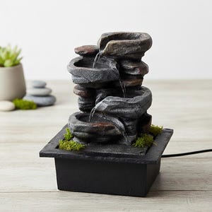 Peut inclure: Une fontaine à eau de table à plusieurs niveaux composée de bassins en pierre grise et brune empilés, avec de l'eau s'écoulant sur chaque étage. La fontaine repose sur une base carrée noire agrémentée de mousse verte et d'un cordon noir.