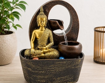May include: A tabletop water fountain featuring a gold-coloured Buddha statue in a meditative pose. The fountain has a dark brown, textured oval frame with a spout that pours water into a small basin. Two small, smooth, colourful stones sit at the base.