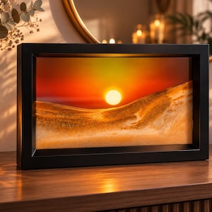 May include: A rectangular black frame containing a dynamic sand art display. The interior features layers of golden and beige sand forming dune-like shapes against a vibrant sunset background with a bright white sun and orange sky.