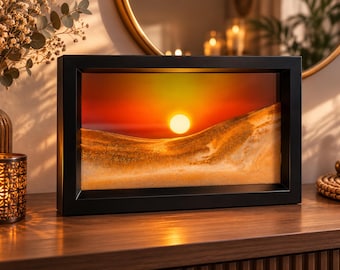 Könnte beinhalten: Ein rechteckiger schwarzer Rahmen mit einem dynamischen Sandbild. Das Innere zeigt Schichten aus goldenem und beigem Sand, die dünenartige Formen vor einem lebendigen Sonnenuntergangshintergrund mit einer hellen weißen Sonne bilden.