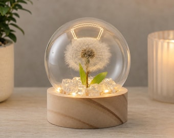 May include: A spherical glass dome containing a single white dandelion seed head and two green leaves, resting on a cylindrical light-coloured wooden base. The base features integrated warm LED lights that illuminate the clear crystals inside.