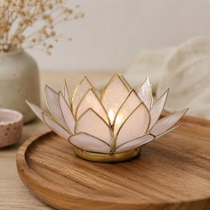 Könnte beinhalten: Ein Lotusblüten-Kerzenhalter mit goldfarbenem Metallrahmen und weißen Blütenblättern. Der Kerzenhalter ist beleuchtet und wirft einen warmen Schein. Der Kerzenhalter steht auf einem Holztablett.