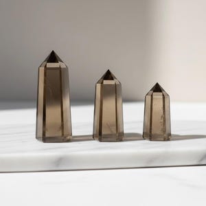 May include: Three smoky quartz crystal towers of varying sizes. The crystals are a translucent brown color with geometric facets and pointed tops. They are arranged on a white marble surface.