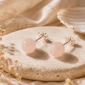 Può includere: Un paio di orecchini a lobo con pietre preziose di quarzo rosa, lisce e rotonde. Ogni pietra è montata su un perno in metallo color argento lucido con una chiusura a farfalla standard, creando un design semplice ed elegante.