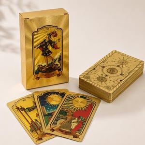 Könnte beinhalten: Ein goldfarbenes Tarot-Deck mit einer vertikalen Schachtel, die die Karte Der Narr zeigt. Drei einzelne Karten liegen offen, darunter Die Sonne, neben einem Kartenstapel mit filigranen goldenen geometrischen und himmlischen Linien.