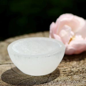 Puede incluir: Un pequeño cuenco blanco translúcido de forma redondeada se encuentra sobre una superficie de madera envejecida. Una flor rosa suave está en el fondo. El cuenco parece estar hecho de un material sólido, posiblemente cristalino.