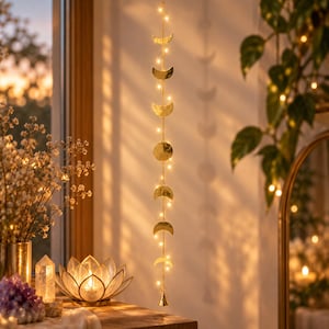 Può includere: Una decorazione verticale sospesa caratterizzata da una serie di fasi lunari in metallo dorato collegate da sottili catenelle, intervallate da piccole luci LED bianco caldo, che termina con un piccolo campanello in metallo alla base.