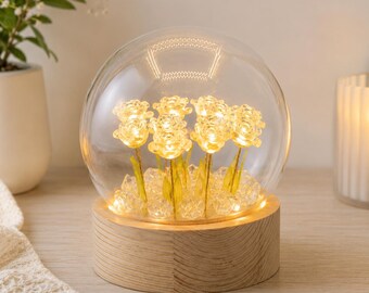 May include: A spherical glass dome lamp featuring a cluster of small, illuminated yellow rose-shaped flowers resting on a circular light-coloured wooden base, surrounded by clear crystal-like decorative stones that reflect the warm internal glow.