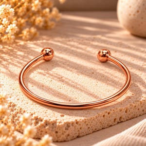 Peut inclure: Un bracelet manchette en métal doré rose poli, composé d'un anneau circulaire fin et lisse se terminant aux deux extrémités par une petite perle sphérique. La surface métallique est très réfléchissante et capte la lumière avec éclat.