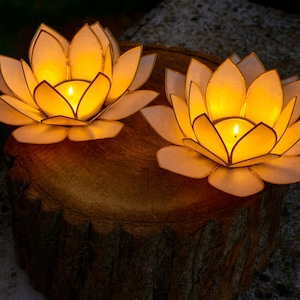 Puede incluir: Dos candelabros iluminados con forma de flor de loto. Los pétalos son de un blanco suave con un brillo amarillo que emana del centro. Los candelabros se asientan sobre una superficie marrón y texturizada, creando un ambiente cálido.