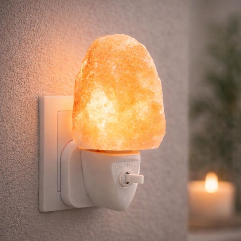 K&ouml;nnte beinhalten: Ein Himalaya-Salz-Nachtlicht, das an eine Steckdose angeschlossen ist. Die Salzlampe hat eine warme orange Farbe und strahlt ein sanftes Leuchten aus. Das Nachtlicht hat einen Ein-/Ausschalter und ist auf einem wei&szlig;en Sockel montiert.