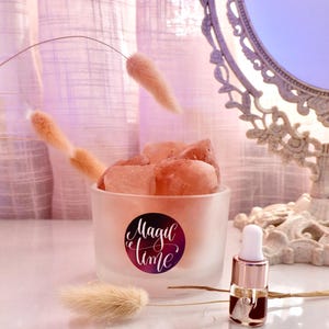 Könnte beinhalten: Ein mattes Glasgefäß mit einem rosa Himalaya-Salzstein darin. Das Gefäß hat einen schwarz-weißen Aufkleber mit der Aufschrift "Magic Time". Neben dem Gefäß befindet sich eine kleine Flasche ätherisches Öl mit goldener Kappe und weißem Tropfer.