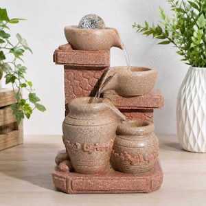 Puede incluir: Una fuente de agua de mesa de varios niveles con un acabado de piedra texturizada de color marrón rojizo. El diseño incluye una esfera de cristal transparente en la parte superior, con agua que cae en cascada a través de cuencos y dos jarrones.