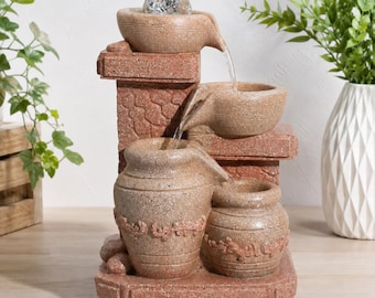May include: A tiered tabletop water fountain featuring a textured, reddish-brown stone finish. The design includes a clear glass sphere at the top, with water cascading down through a series of stacked bowls and two decorative jars at the base.