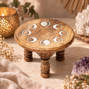 Pode incluir: Um suporte de pedestal redondo de madeira com três pernas esculpidas. A superfície superior apresenta uma disposição circular de oito fases lunares pintadas de branco, incluindo crescentes e círculos cheios, sobre madeira castanha.